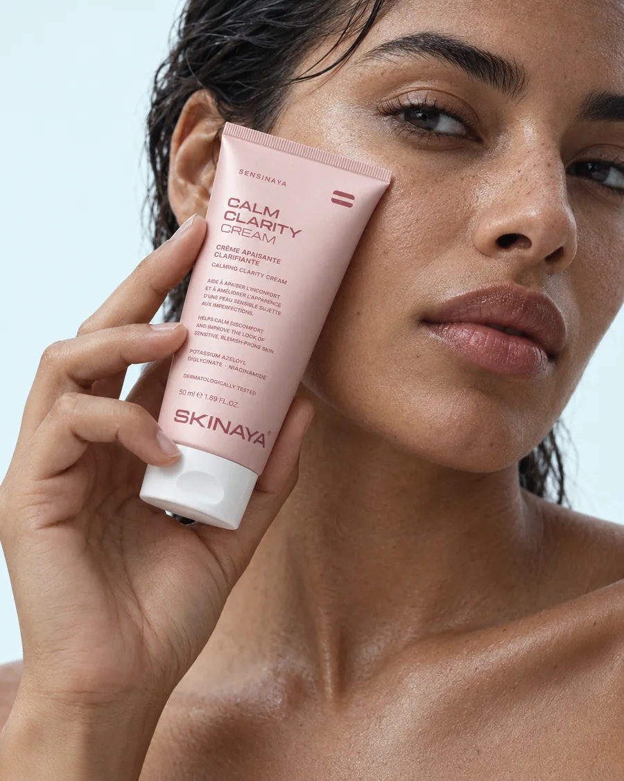 A woman holding the SENSINAYA Calm Clarity Cream tube against her cheek