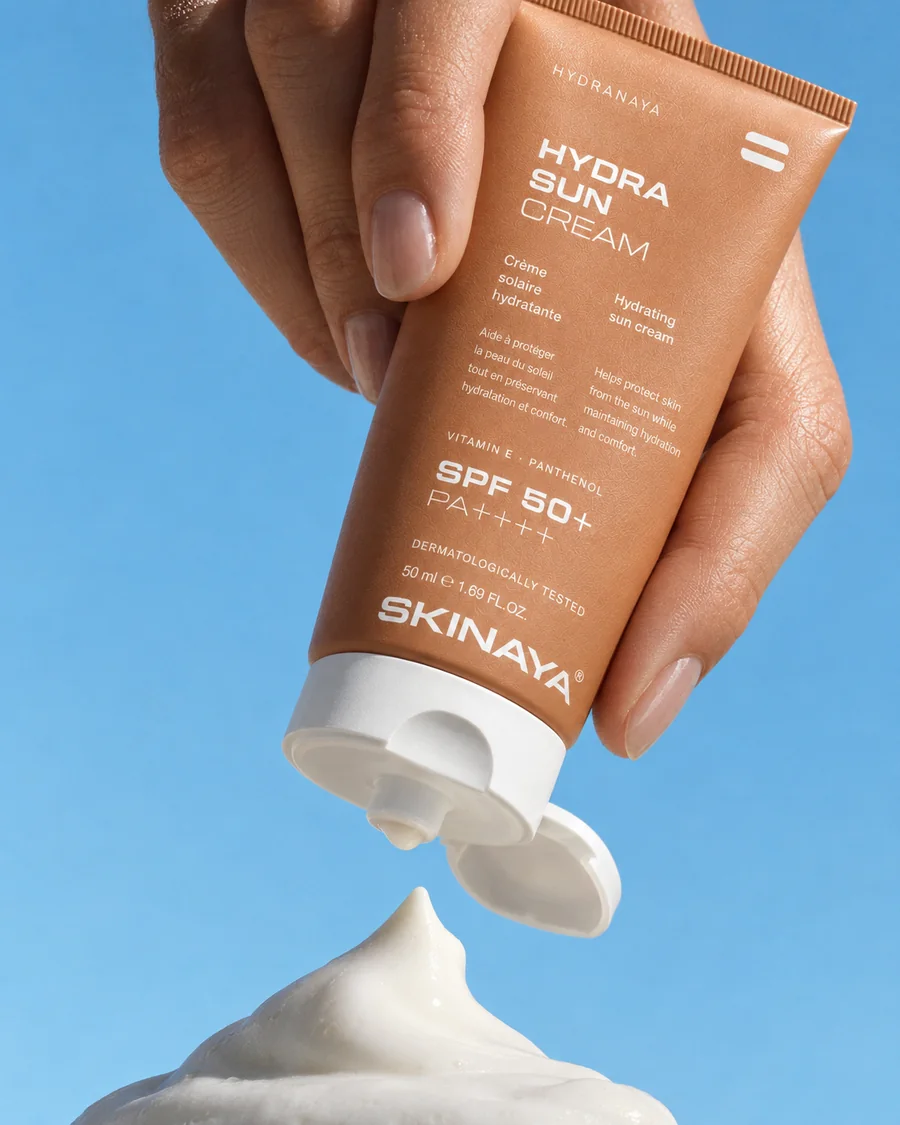 A hand holding the HYDRANAYA Hydra Sun Cream above a dollop of cream against a clear sky blue