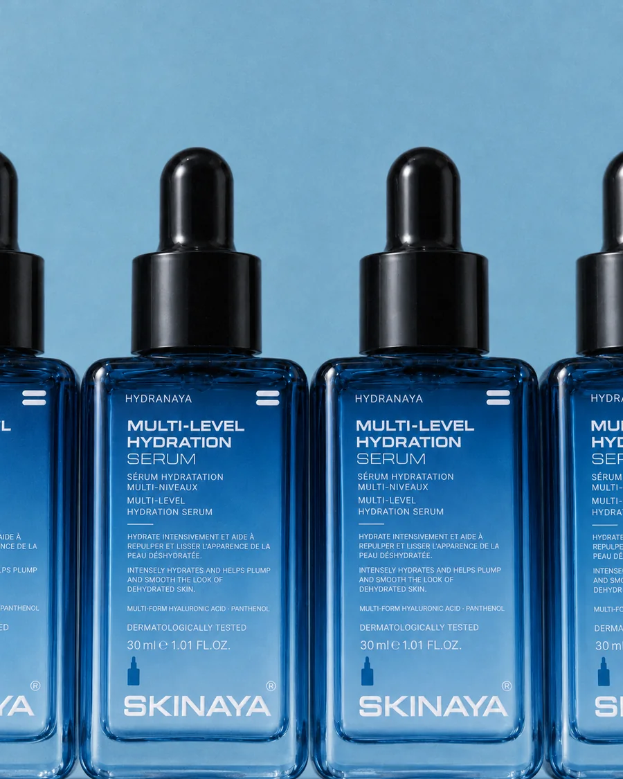 A row of identical HYDRANAYA Multi-Level Hydration Serum bottles on dusty sky blue
